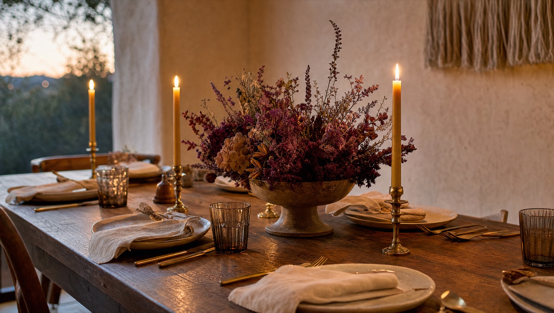 A long wooden table at dusk set with lit beeswax candles and a low bowl of deep red and plum dried flowers.