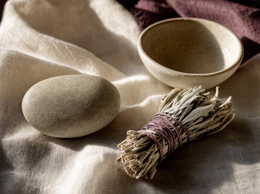 A smooth stone and a bundle of dried sage tied with plum thread, resting on cream linen in low morning light.